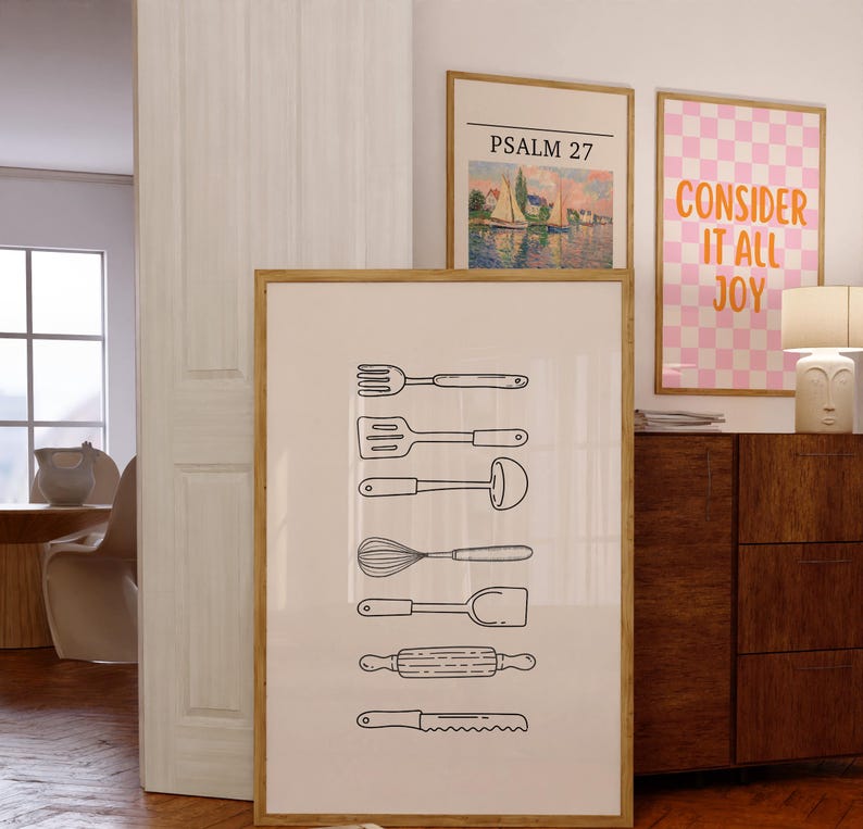 May include: A framed print featuring line drawings of kitchen utensils, including a fork, spatula, ladle, whisk, spoon, rolling pin, and knife. The print is in a wooden frame and is displayed with other framed art.