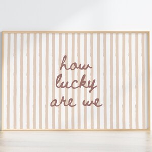 May include: A framed print with a light wood frame. The background features vertical stripes in beige and white. The text "how lucky are we" is written in a cursive, mauve font.