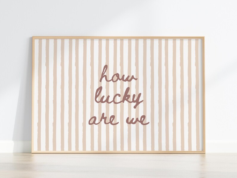 May include: A framed print with a light wood frame. The background features vertical stripes in beige and white. The text "how lucky are we" is written in a cursive, mauve font.