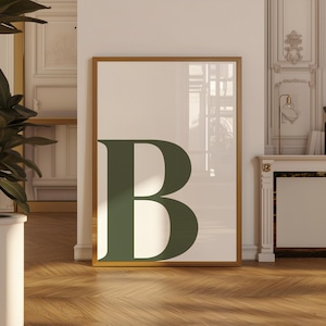 May include: A framed art print featuring a large, dark green letter "B" against a cream-colored background. The artwork is encased in a gold-colored frame, creating a modern and minimalist aesthetic. The print is displayed on a wooden floor.