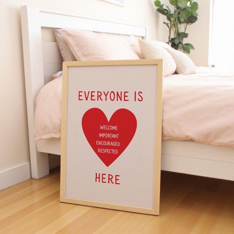 May include: A framed print with the words "EVERYONE IS" above a red heart shape. Inside the heart are the words "WELCOME IMPORTANT ENCOURAGED RESPECTED". Below the heart, the word "HERE" is printed in red. The frame is light wood.