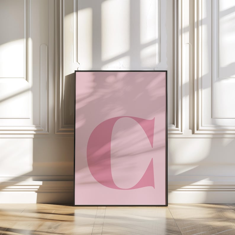 Puede incluir: Un p&oacute;ster rosa con una gran letra "C" rosa y en negrita en el centro. El p&oacute;ster est&aacute; enmarcado en un marco negro y est&aacute; apoyado contra una pared blanca.