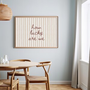 May include: A framed wall art piece with a beige and white striped background and the text "how lucky are we" in a cursive font. The artwork is displayed in a dining room setting with a wooden table and chairs.