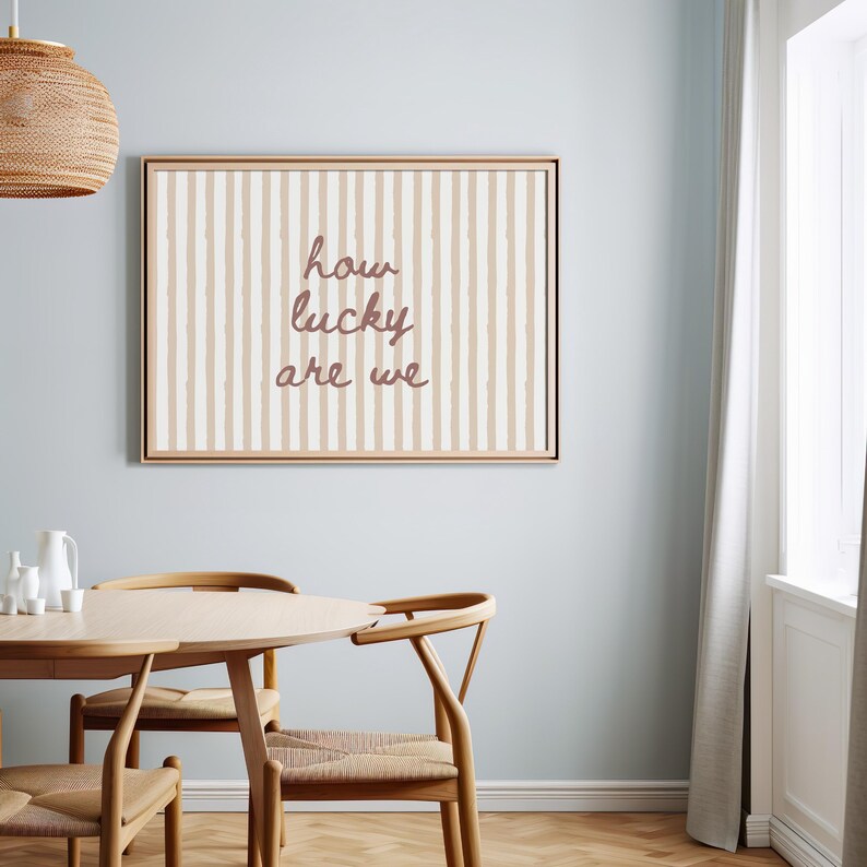 May include: A framed wall art piece with a beige and white striped background and the text "how lucky are we" in a cursive font. The artwork is displayed in a dining room setting with a wooden table and chairs.