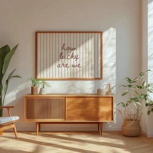 May include: A mid-century modern living room with a wooden cabinet, a framed artwork with the text "how lucky are we", and various plants. The room has a neutral color palette with natural light streaming in from a window.