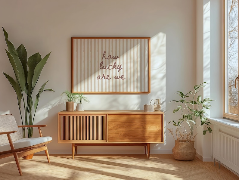 May include: A mid-century modern living room with a wooden cabinet, a framed artwork with the text "how lucky are we", and various plants. The room has a neutral color palette with natural light streaming in from a window.