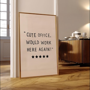 May include: A framed print with the text "CUTE OFFICE, WOULD WORK HERE AGAIN!" in black font. The print has a gold frame and five black star icons. The print is leaning against a white door frame on a wooden floor.