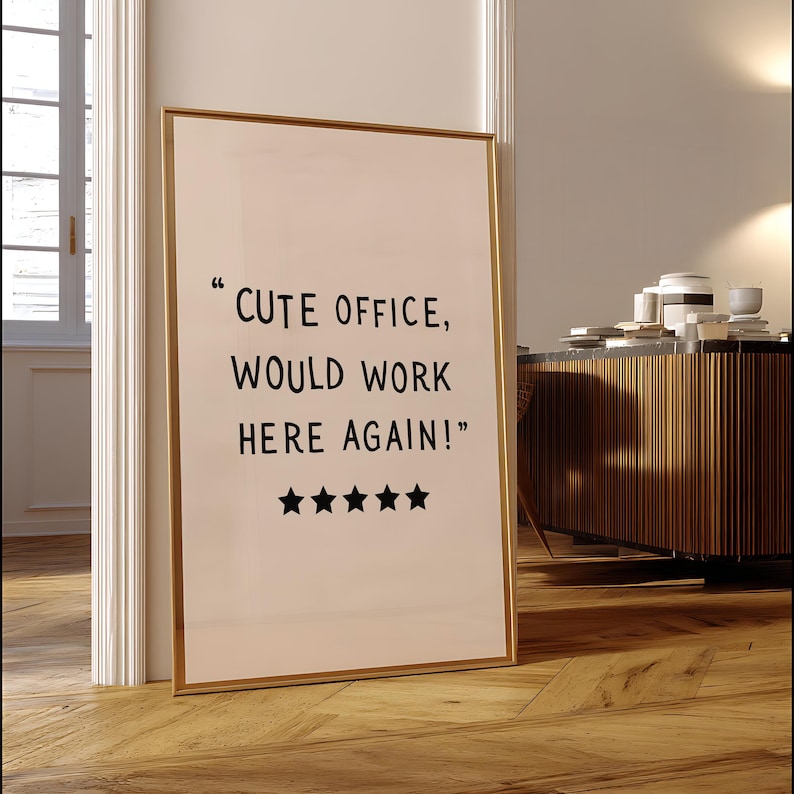 May include: A framed print with the text "CUTE OFFICE, WOULD WORK HERE AGAIN!" in black font. The print has a gold frame and five black star icons. The print is leaning against a white door frame on a wooden floor.