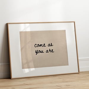 May include: A framed print with the words "come as you are" in black script on a beige background. The frame is a light brown color and is leaning against a white wall on a wooden floor.