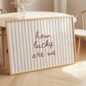 May include: A framed print with a beige and white striped pattern and the words "how lucky are we" in a handwritten style. The print is in a light wood frame, leaning against a table with chairs.
