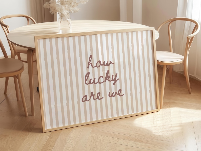 May include: A framed print with a beige and white striped pattern and the words "how lucky are we" in a handwritten style. The print is in a light wood frame, leaning against a table with chairs.