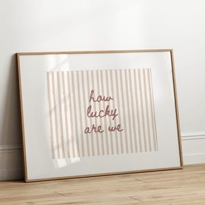 May include: Framed art print with a light brown wooden frame. The artwork features a striped background in beige and white, with the phrase "how lucky are we" in a mauve script font. The print is leaning against a white wall on a wooden floor.