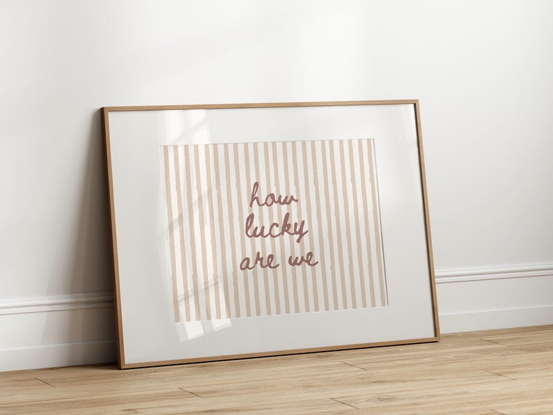 May include: Framed art print with a light brown wooden frame. The artwork features a striped background in beige and white, with the phrase "how lucky are we" in a mauve script font. The print is leaning against a white wall on a wooden floor.