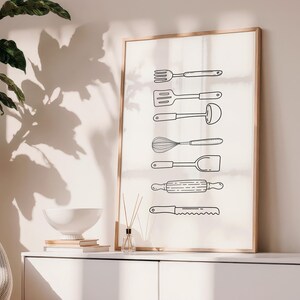 May include: A framed print featuring line drawings of kitchen utensils, including a fork, spatula, ladle, whisk, turner, rolling pin, and serrated knife. The artwork is displayed on a white cabinet with a plant and decorative items.