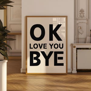 Pode incluir: Uma impressão de arte emoldurada com as palavras "OK LOVE YOU BYE" em letras pretas em negrito sobre um fundo creme. A impressão está em uma moldura dourada e encostada em uma parede branca. O chão é de madeira.