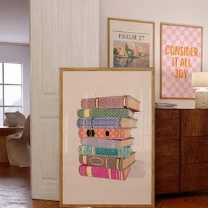 May include: A framed print of a stack of colorful, patterned books with a light beige background. The print is in a wooden frame. The text "PSALM 27" is visible in a smaller framed print above the book print.
