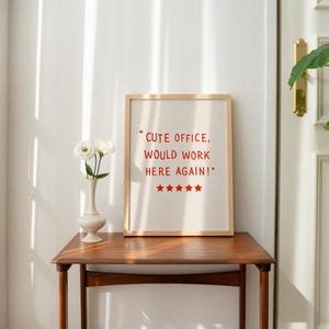 May include: A framed print with the text "Cute office, would work here again!" in red. The print is on a wooden table, next to a vase with white flowers. The room has a white door and a plant.