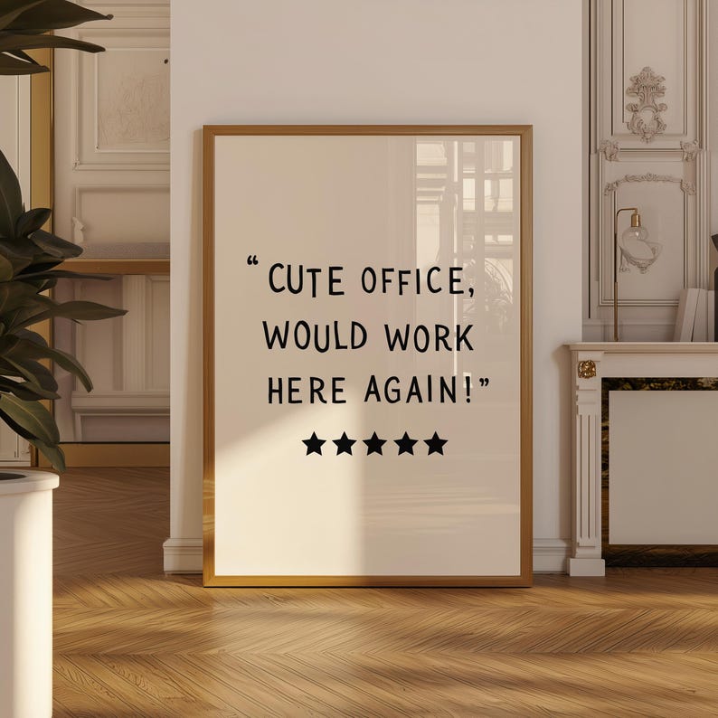 May include: A framed print with a gold frame and the text "CUTE OFFICE, WOULD WORK HERE AGAIN!" in black. Below the text are five black stars. The print is displayed in a room with a wooden floor.