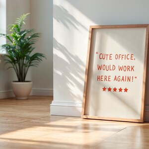 May include: A framed print with the text "CUTE OFFICE, WOULD WORK HERE AGAIN!" in red. The print is leaning against a white wall. A potted plant and a window are visible in the background.