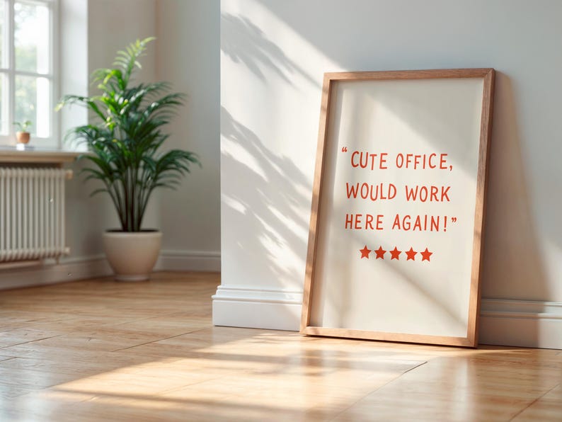 May include: A framed print with the text "CUTE OFFICE, WOULD WORK HERE AGAIN!" in red. The print is leaning against a white wall. A potted plant and a window are visible in the background.
