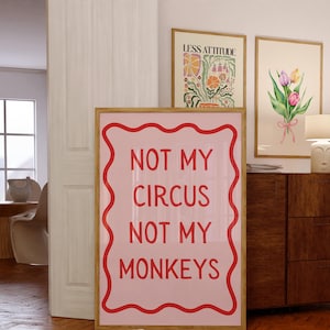 May include: A framed pink poster with the text "NOT MY CIRCUS NOT MY MONKEYS" in red. The poster is in a room with other framed art and a wooden cabinet. The room has a neutral color palette.