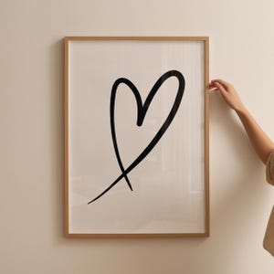 May include: A framed print featuring a large, black heart drawn with a single, continuous line. The artwork is set against a white background and encased in a light brown wooden frame. The image is mounted on a neutral-colored wall.