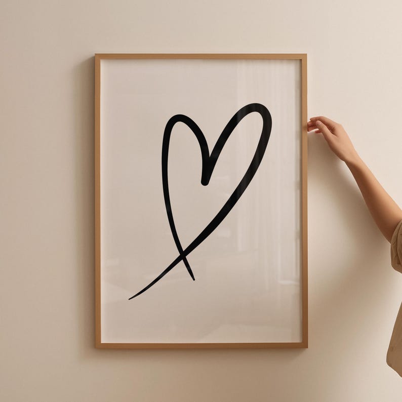 May include: A framed print featuring a large, black heart drawn with a single, continuous line. The artwork is set against a white background and encased in a light brown wooden frame. The image is mounted on a neutral-colored wall.