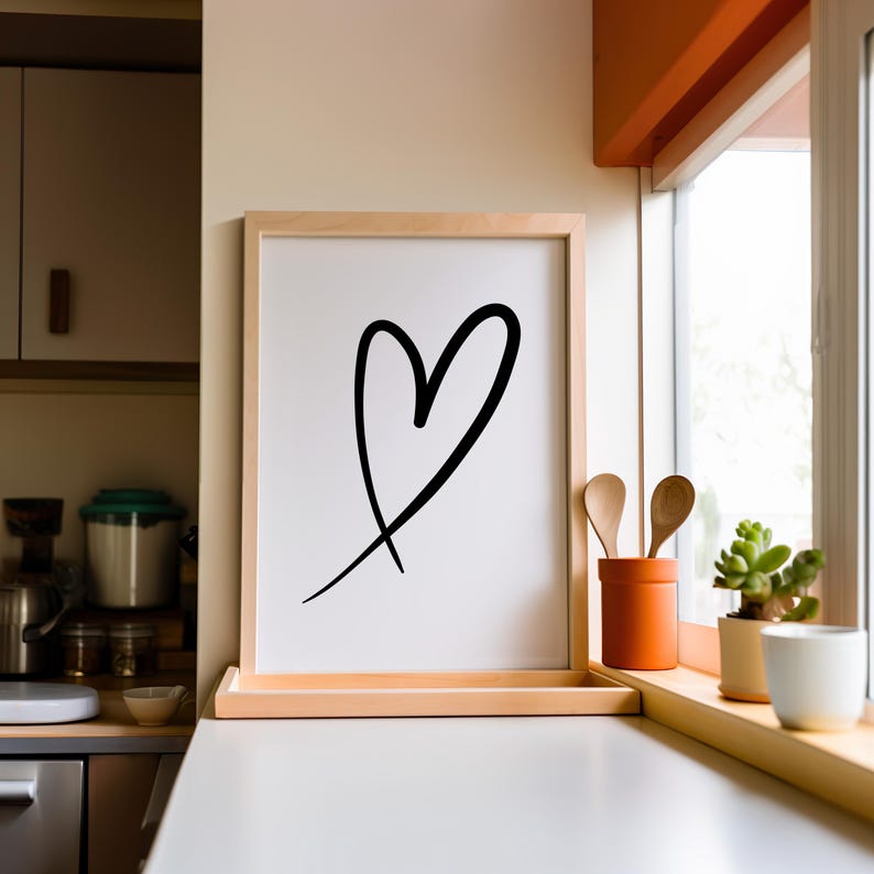 May include: A framed print featuring a black heart outline on a white background. The artwork is displayed in a light wood frame, resting on a white countertop. The kitchen setting includes a window, wooden spoons, and a small potted plant.