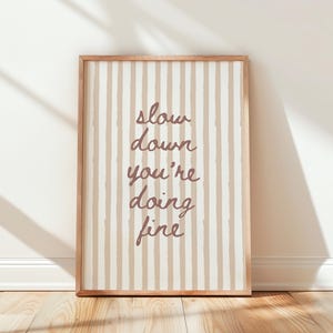 Puede incluir: Una impresión enmarcada con un marco de madera clara. La obra de arte presenta rayas verticales en beige y blanco. Las palabras "slow down you're doing fine" están escritas en una fuente cursiva en color marrón apagado.