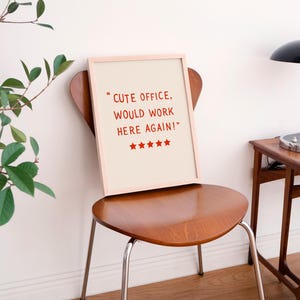 May include: A framed print with the text "CUTE OFFICE, WOULD WORK HERE AGAIN!" in red, with red stars below. The print rests on a wooden chair with metal legs. A black desk lamp and small wooden table are in the background.