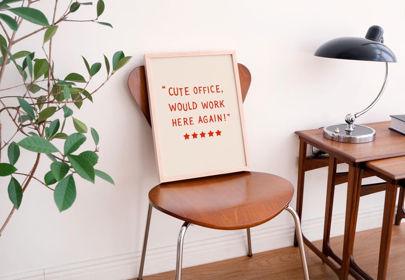 May include: A framed print with the text "CUTE OFFICE, WOULD WORK HERE AGAIN!" in red, with red stars below. The print rests on a wooden chair with metal legs. A black desk lamp and small wooden table are in the background.