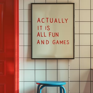 May include: A framed print with the text "ACTUALLY IT IS ALL FUN AND GAMES" in red letters. The print hangs on a white tiled wall. A blue stool sits below the print, and a red door is visible on the left.