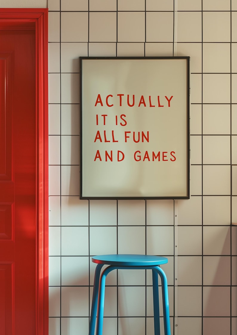 May include: A framed print with the text "ACTUALLY IT IS ALL FUN AND GAMES" in red letters. The print hangs on a white tiled wall. A blue stool sits below the print, and a red door is visible on the left.