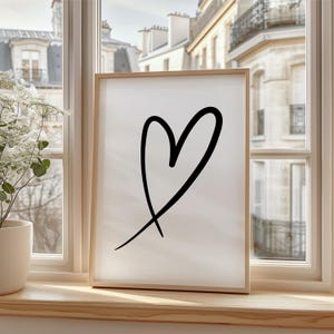 May include: A framed art print featuring a black heart design on a white background. The artwork is displayed in a light wood frame and is positioned on a wooden windowsill. The window overlooks a European-style cityscape.