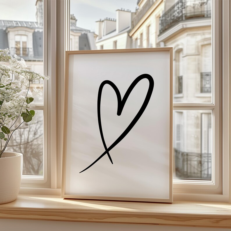 May include: A framed art print featuring a black heart design on a white background. The artwork is displayed in a light wood frame and is positioned on a wooden windowsill. The window overlooks a European-style cityscape.