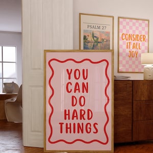 Peut inclure: Trois impressions d'art encadrées avec des citations inspirantes et des scènes de paysage. La plus grande impression a un fond rose avec du texte rouge qui dit "YOU CAN DO HARD THINGS." Les autres impressions présentent une scène de voilier et un motif à carreaux roses et blancs.