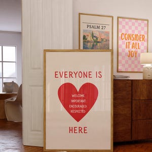 以下が含まれることがあります： 赤いハートの形の上に「EVERYONE IS」という言葉が書かれた額入りのポスター。ハートの中には「WELCOME, IMPORTANT, ENCOURAGED, RESPECTED」という言葉が書かれています。ハートの下には「HERE」という言葉が印刷されています。他の2つの額入りのポスターが見えます。