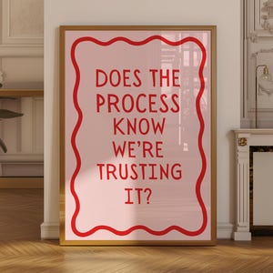 Op de afbeelding: Een ingelijste kunstdruk met een lichtroze achtergrond en rode tekst die luidt: "DOES THE PROCESS KNOW WE'RE TRUSTING IT?" De tekst staat in een rode, golvende rand. De lijst is lichtbruin.