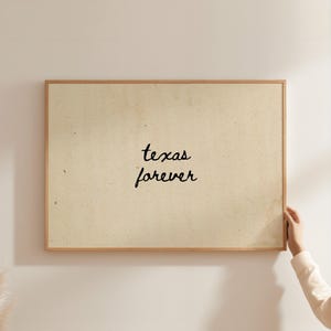 Può includere: Un'opera d'arte da parete rettangolare con uno sfondo in carta testurizzata beige chiaro e il testo nero scritto a mano "texas forever" centrato al centro. L'opera è racchiusa in una sottile cornice in legno di colore chiaro.