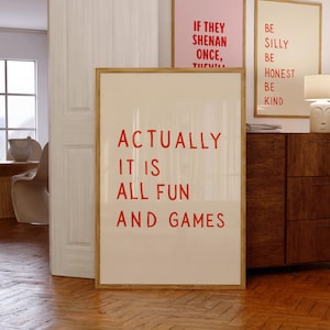 Puede incluir: Un cuadro rectangular con marco, fondo beige claro y texto rojo en negrita que dice: ACTUALLY IT IS ALL FUN AND GAMES. El marco es de madera de color claro y la lámina se encuentra en una habitación con suelo de madera.