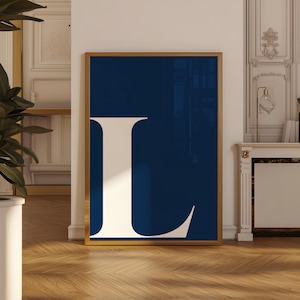 May include: A framed art print featuring a large, stylized letter "L" in a cream color against a deep navy blue background. The artwork is displayed in a gold-colored frame, adding a touch of elegance to the design. The print is displayed on a wooden floor.