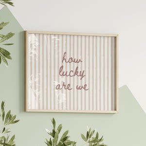 May include: A framed print with the words "how lucky are we" in a cursive font. The background has vertical beige and white stripes. The frame is light wood, and the print is hanging on a wall with green and white paint.