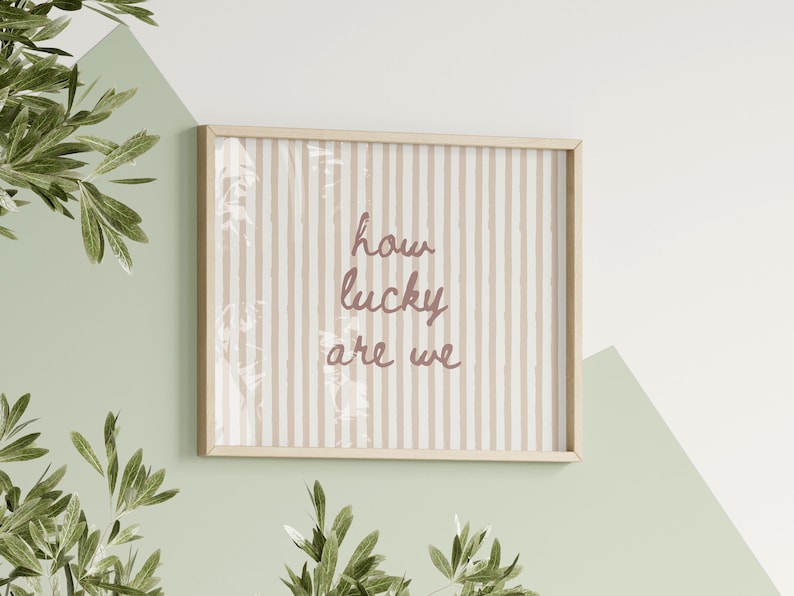 May include: A framed print with the words "how lucky are we" in a cursive font. The background has vertical beige and white stripes. The frame is light wood, and the print is hanging on a wall with green and white paint.