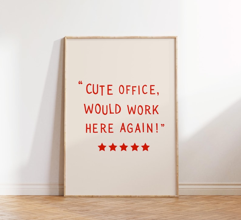 May include: A framed print with the text "CUTE OFFICE, WOULD WORK HERE AGAIN!" in red, along with five red stars. The print has a cream-colored background and a light wooden frame, leaning against a white wall.
