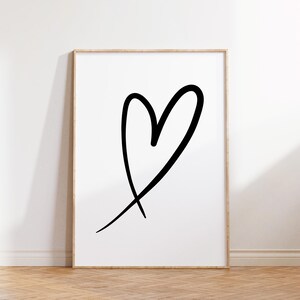 May include: A framed art print featuring a large, black, hand-drawn heart on a white background. The heart is a simple, bold outline with a slightly uneven shape, giving it a casual, artistic feel. The frame is a light wood color.