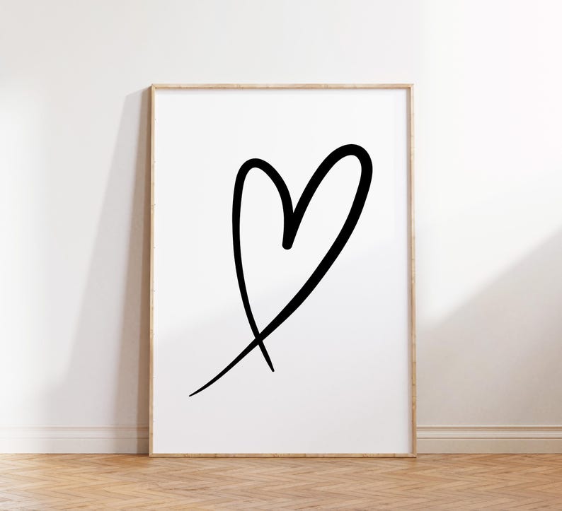 May include: A framed art print featuring a large, black, hand-drawn heart on a white background. The heart is a simple, bold outline with a slightly uneven shape, giving it a casual, artistic feel. The frame is a light wood color.