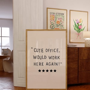 May include: A framed print with the text "CUTE OFFICE, WOULD WORK HERE AGAIN!" and five black stars. Two other framed art pieces are visible, one with floral art and the other with the text "LESS ATTITUDE". A wooden cabinet and a lamp are also in the image.