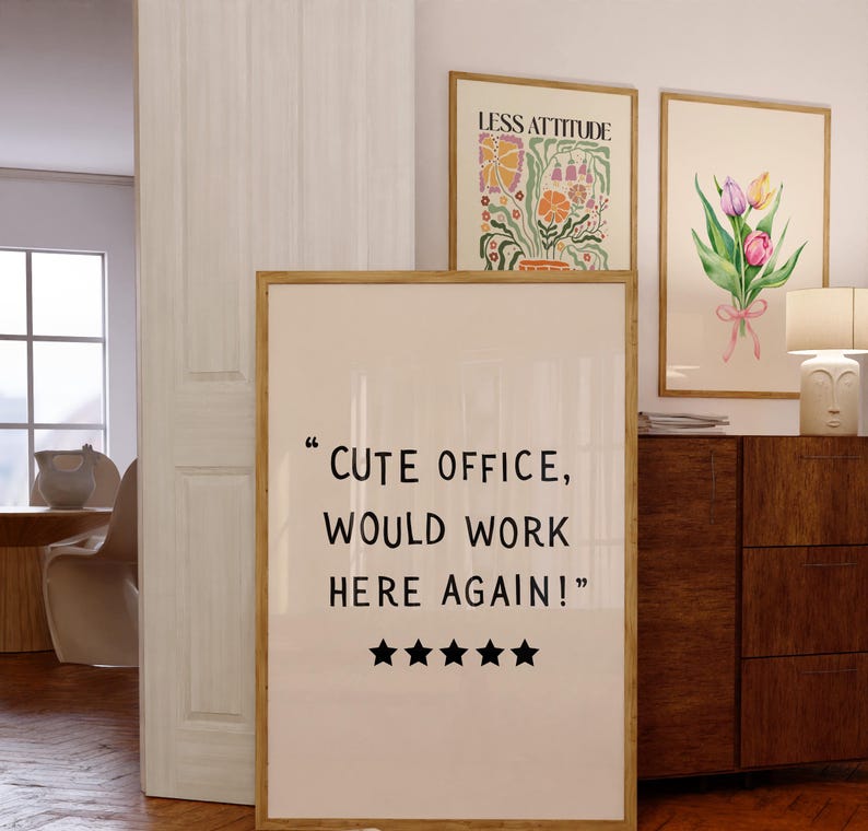 May include: A framed print with the text "CUTE OFFICE, WOULD WORK HERE AGAIN!" and five black stars. Two other framed art pieces are visible, one with floral art and the other with the text "LESS ATTITUDE". A wooden cabinet and a lamp are also in the image.
