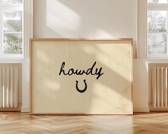 May include: A framed print with a vintage-style background. The black text says "howdy" above a black horseshoe. The frame is a light brown color. The print is displayed on a wooden floor.