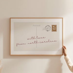 Puede incluir: Una lámina de arte mural enmarcada con un diseño de postal en color beige que incluye el texto "Postcard" en la parte superior y "with love from north carolina" en letra cursiva. Contiene un matasellos circular y un sello rectangular floral.